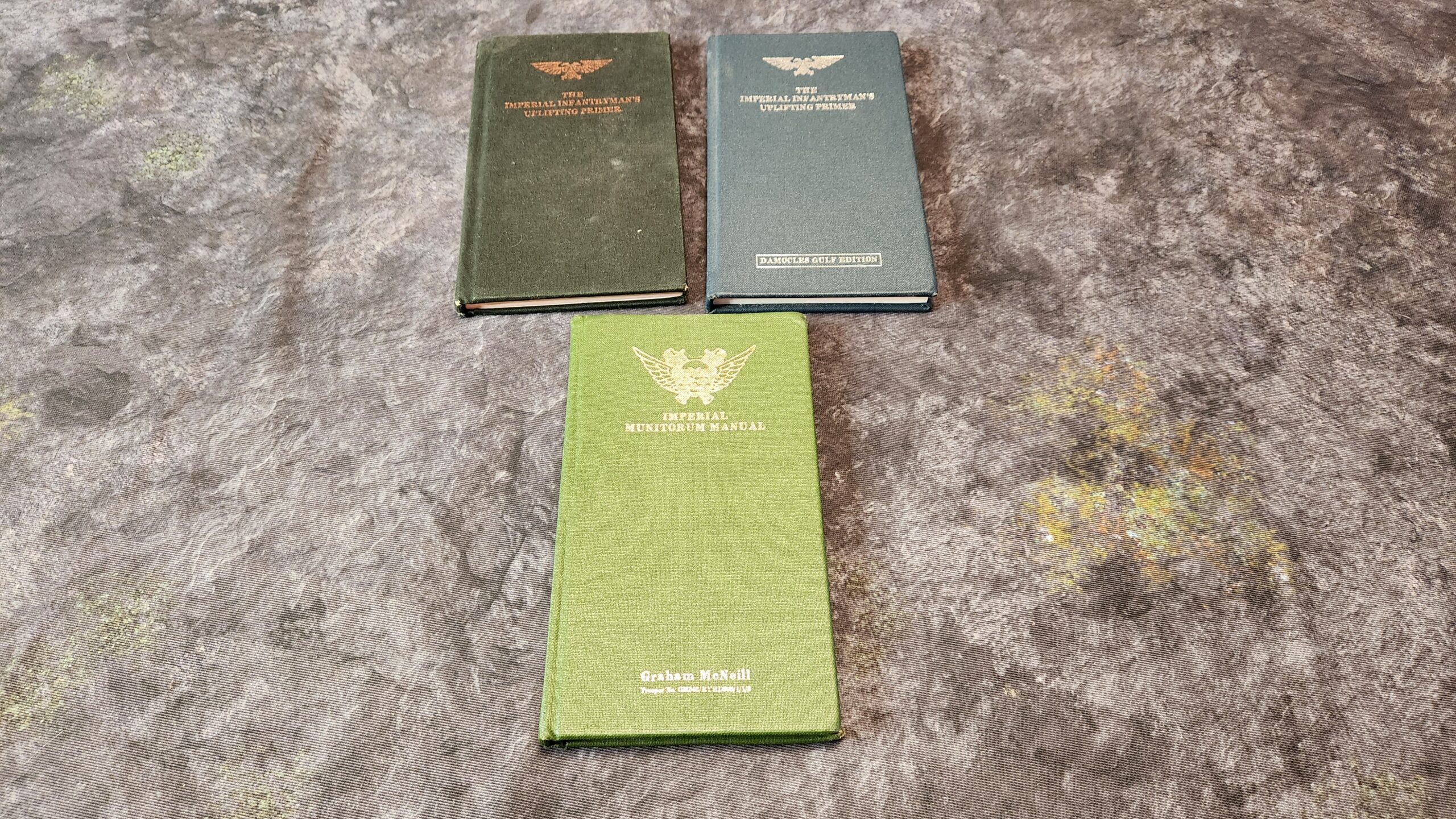 A picture of three books: The Imperial Infantryman's Uplifting Primer, the Imperial Infantryman's Uplifting Primer Damocles Gulf Edition, and the Imperial Munitorum Manual