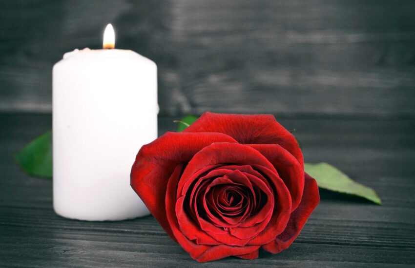 A white candle, lit, next to a red rose, on a grey wooden table.